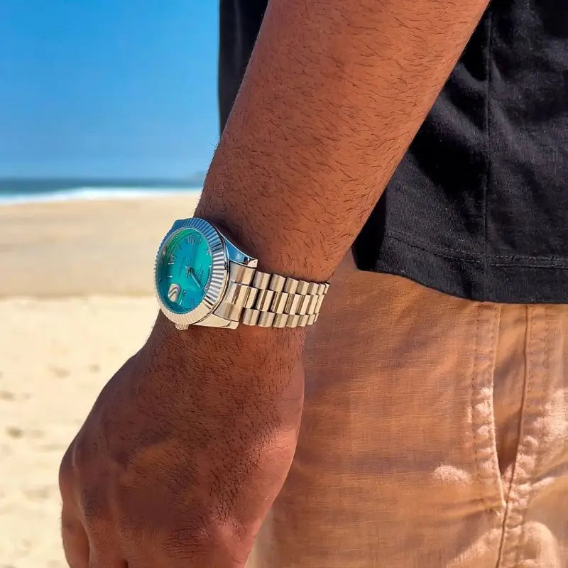 VEXO Day Date wristwatch with turquoise dial on wrist at the beach, silver bracelet and fluted bezel in sunlight